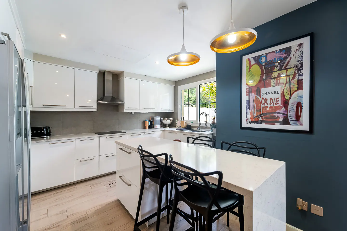 Bright kitchen with white cabinets, island with black stools, and modern pendant lights. A colorful "Chanel or Die" art piece hangs on a blue wall.
