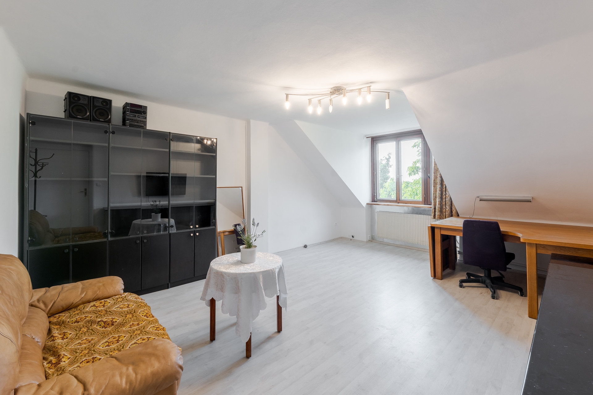 Attic room with white walls, light wood floors, a tan leather sofa, a small round table, and a wooden desk with a chair. [AI-generated text]