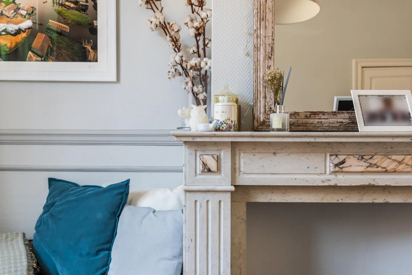 Living room with a marble fireplace, decorated with cotton flowers, a candle, and a framed picture. Blue pillows on a sofa.