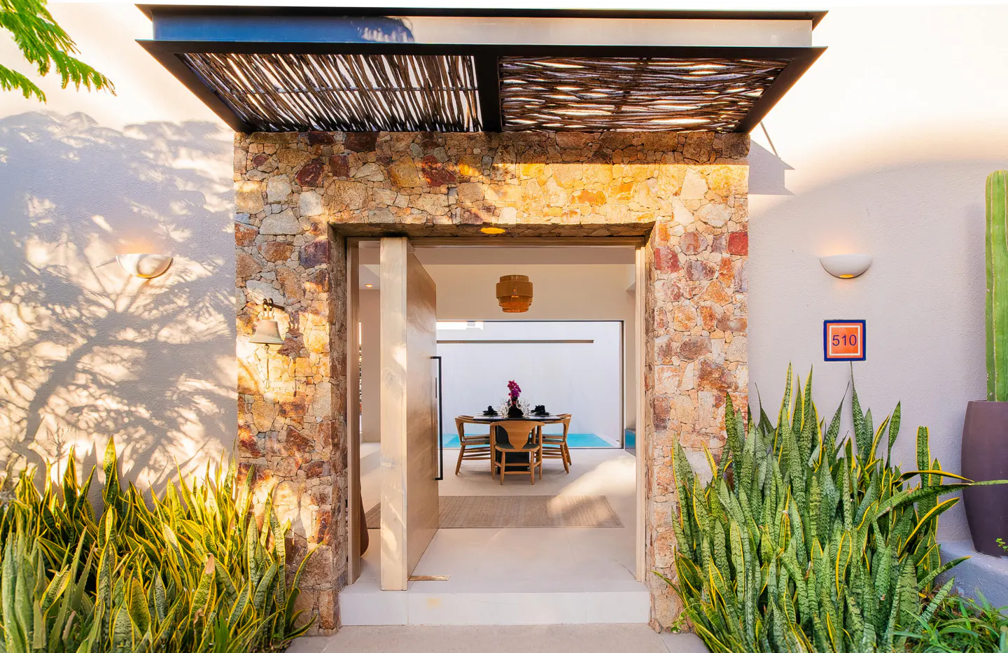 Open doorway framed by stone walls and green plants, leading to a dining area with a pool view.