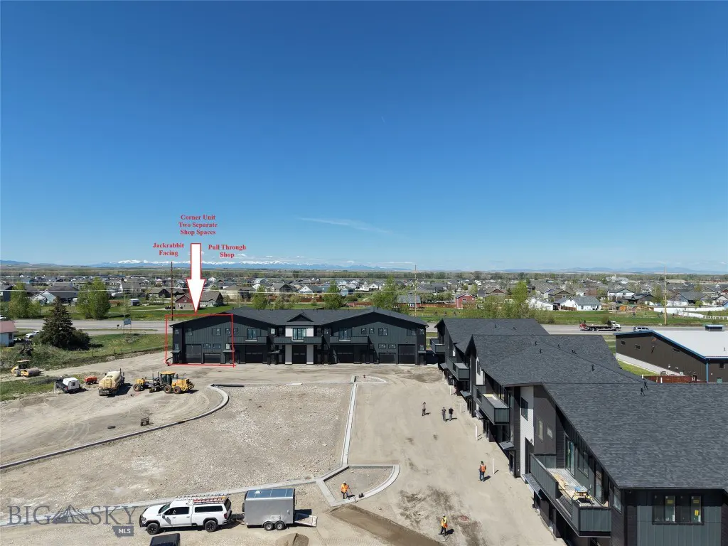 Aerial view of a modern, dark gray commercial building with a flat, gravel lot and construction equipment. Blue sky and distant mountains in the background.