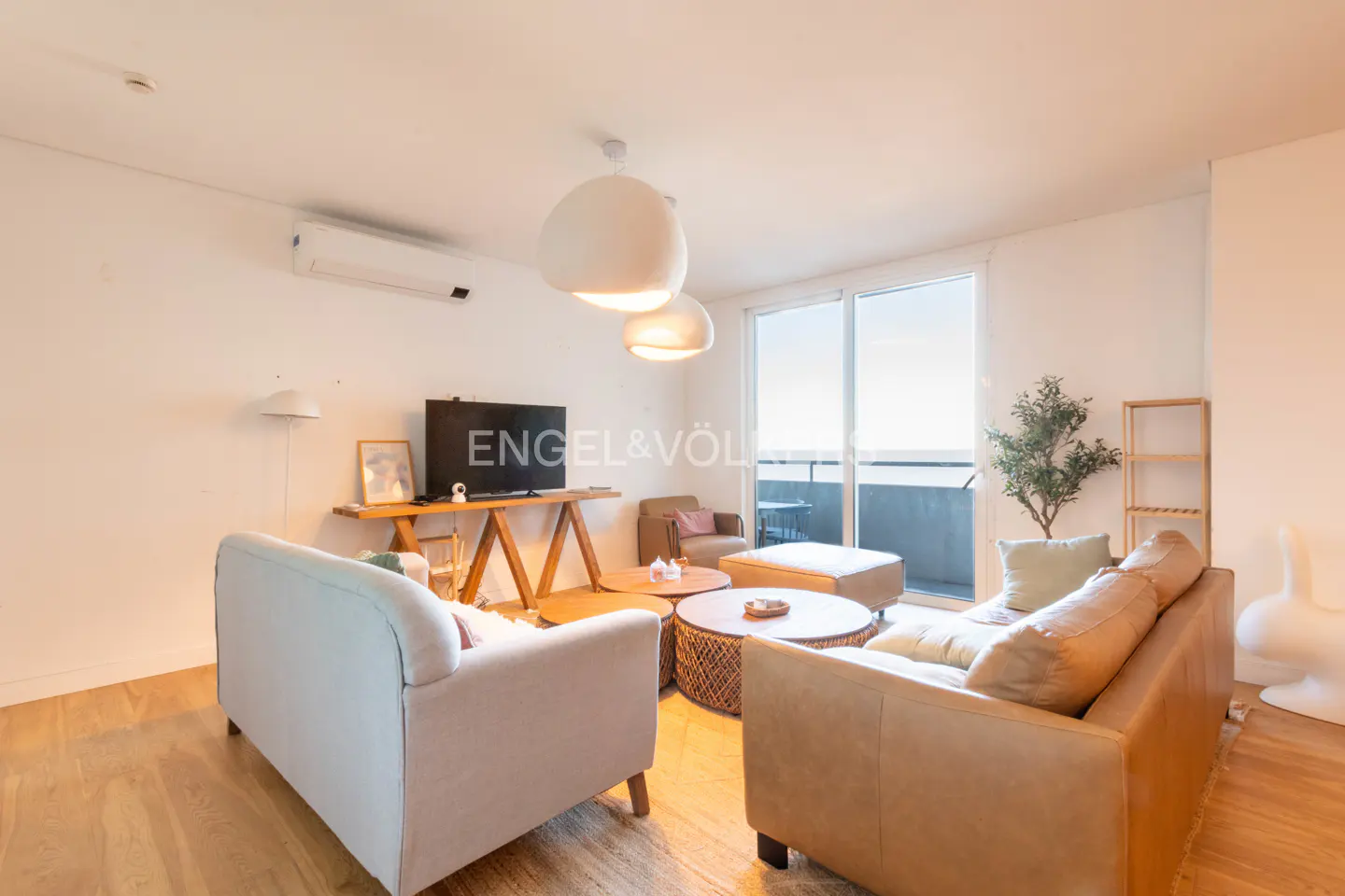 Bright living room with light wood floors, white walls, and tan and white sofas. A TV sits on a wooden table. Balcony with ocean view.
