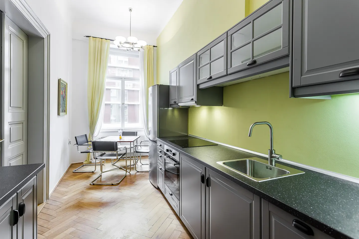 Bright kitchen with gray cabinets, black countertops, and stainless steel appliances. Table and chairs near a window with yellow curtains.