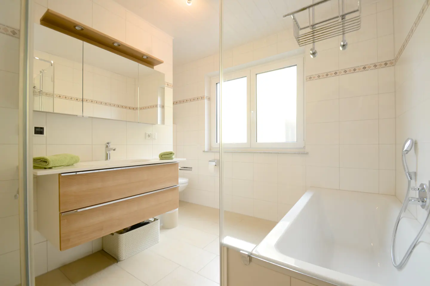 Bright bathroom with white tile, a wood vanity with a white sink, and a white bathtub. A window provides natural light.