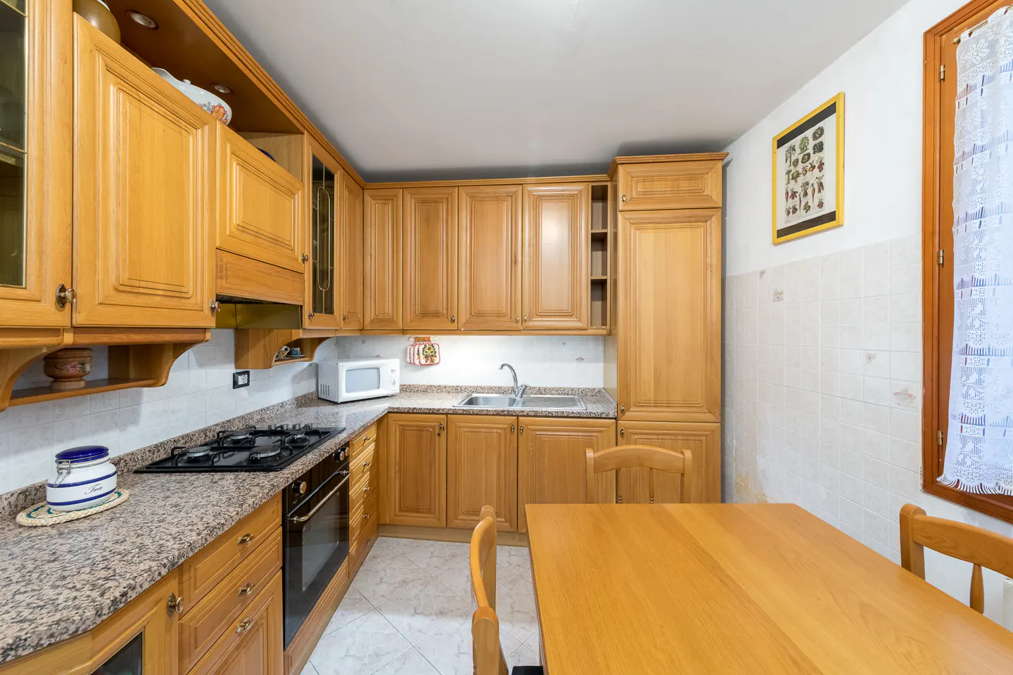 A kitchen with light wood cabinets, granite countertops, and a wooden table with chairs. A window with a white curtain is on the right.