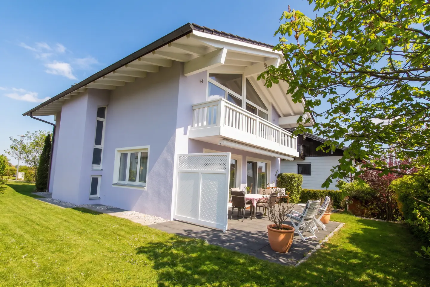 Two-story lilac house with a white balcony and trim, a patio with furniture, and a green lawn.