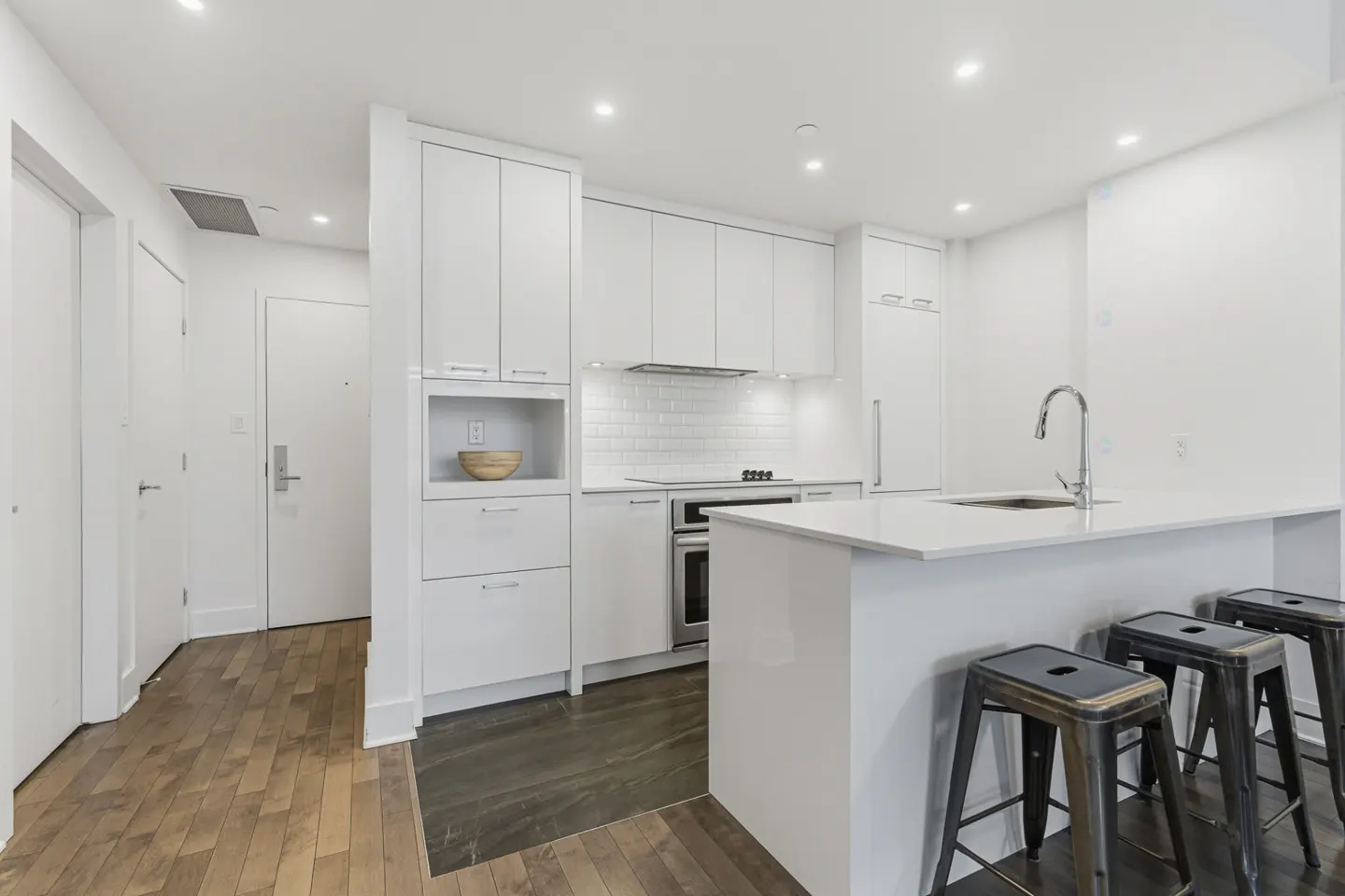 Bright, modern kitchen with white cabinets, countertops, and subway tile backsplash. Three metal stools line the island. Wood floors transition to dark tile.