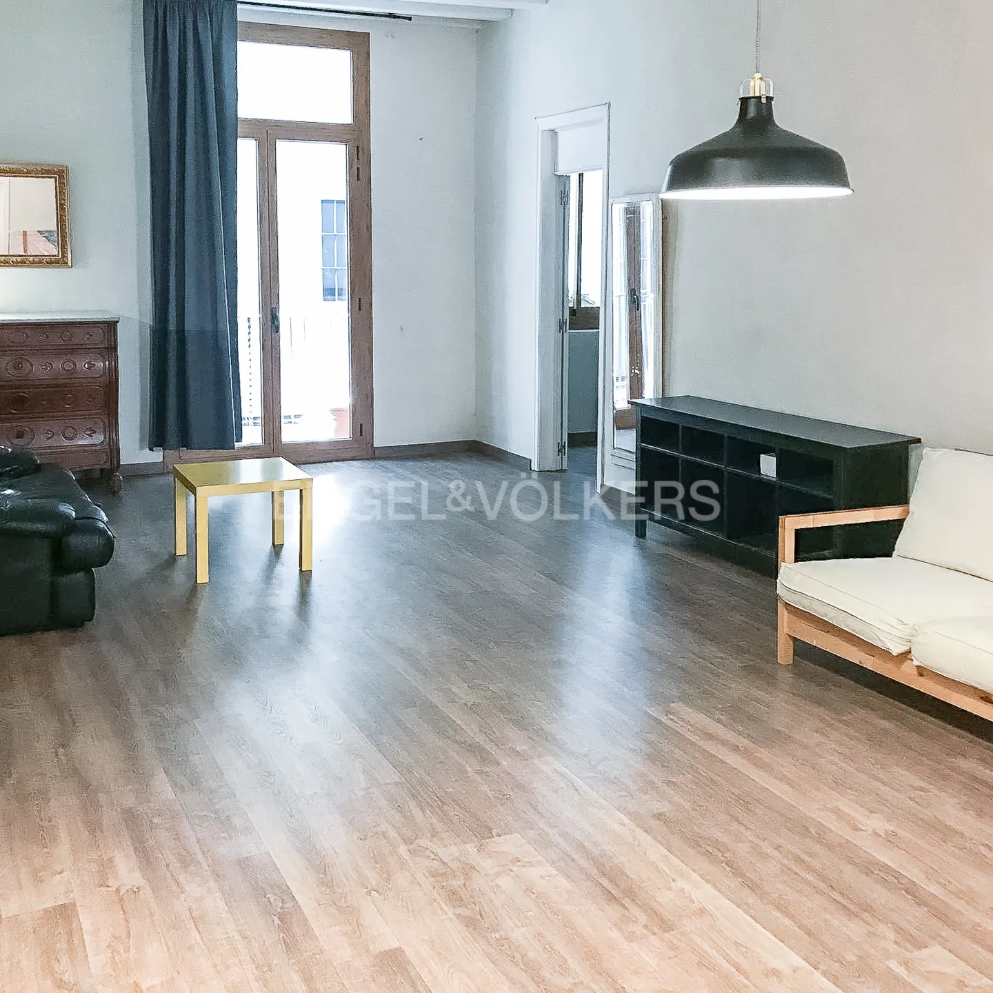 Bright, spacious living room with wood floors, white walls, and a black pendant light. A sofa, table, and dresser furnish the room.