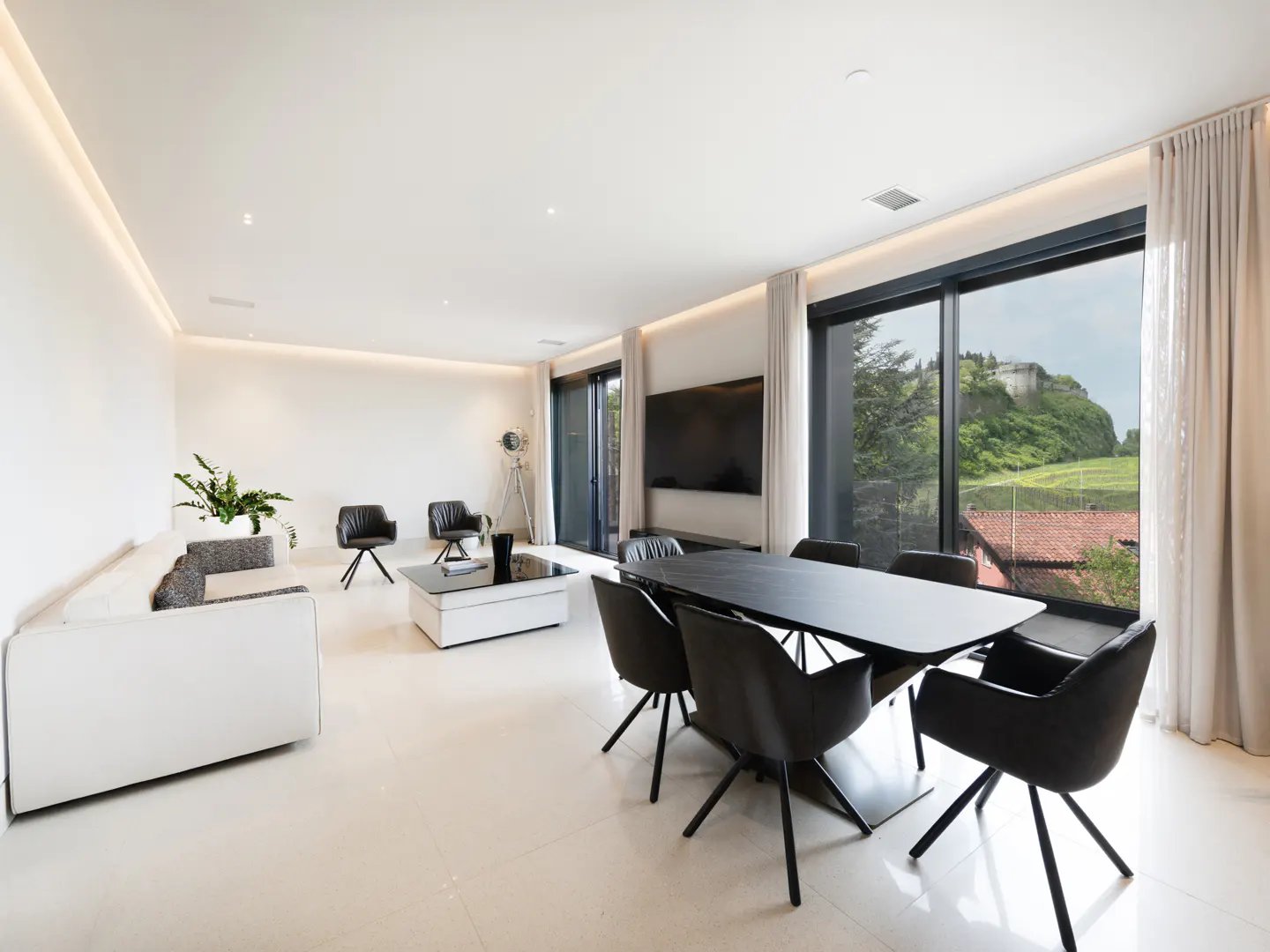 Bright, modern living and dining area with white walls, tile floors, and black dining set. Large windows offer a view of a castle on a hill.