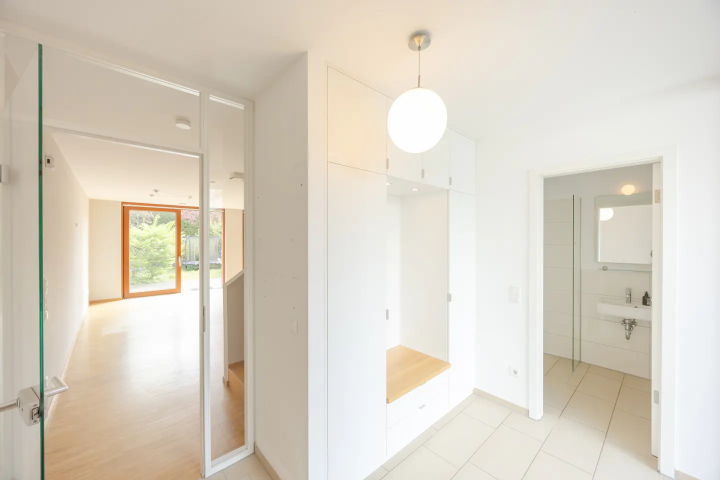 Bright, modern home entryway with white walls, tile floor, and built-in storage. A glass door leads to a living area with a view of the garden.