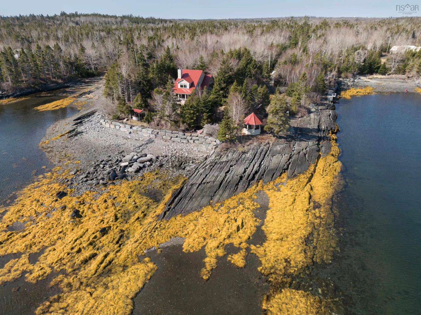 Aerial view of a red-roofed house on a rocky island with a gazebo, surrounded by trees and water.