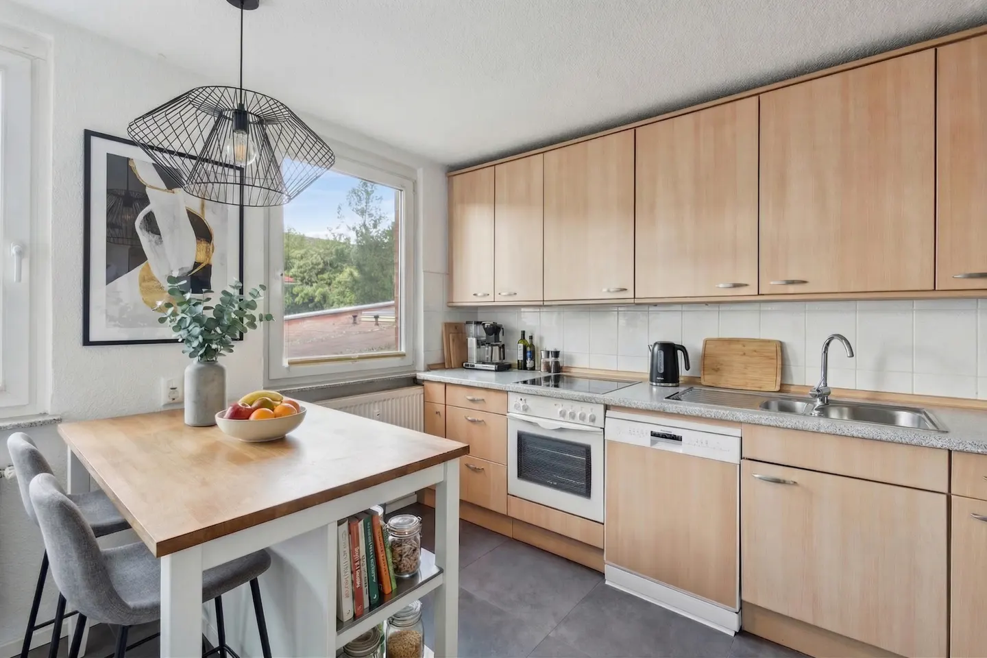 Bright kitchen with light wood cabinets, white appliances, and a gray floor. A wooden island with gray stools sits near a window.