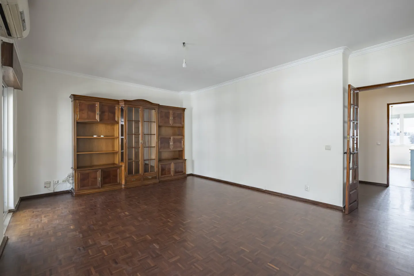 An empty living room with parquet floors, white walls, and a large wooden bookcase. A doorway leads to another room.