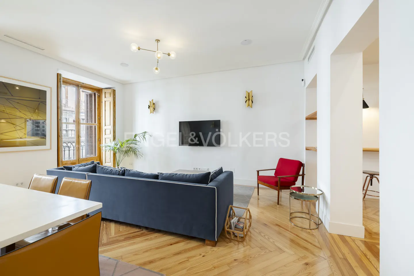 Bright living room with blue sofa, red chair, and herringbone wood floors. Large window with shutters and a modern light fixture.