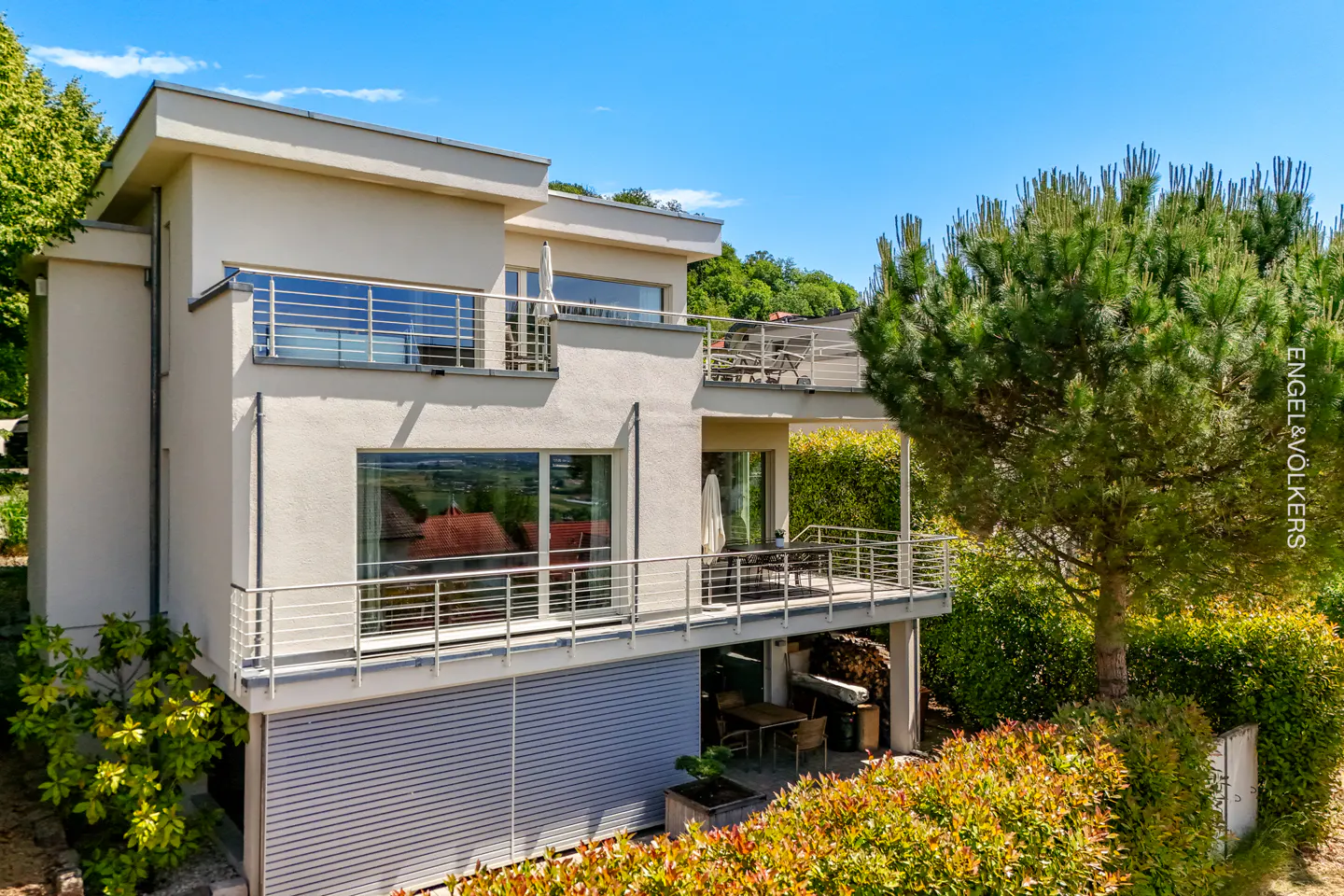 Modern beige two-story house with balconies, large windows, and a lower-level garage, surrounded by lush green trees.