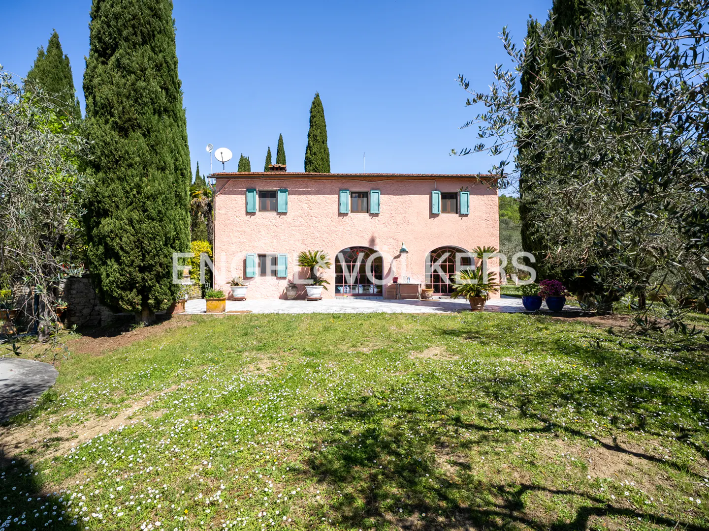 Exterior view of a two-story pink house with turquoise shutters, surrounded by green trees and a lawn with white flowers.