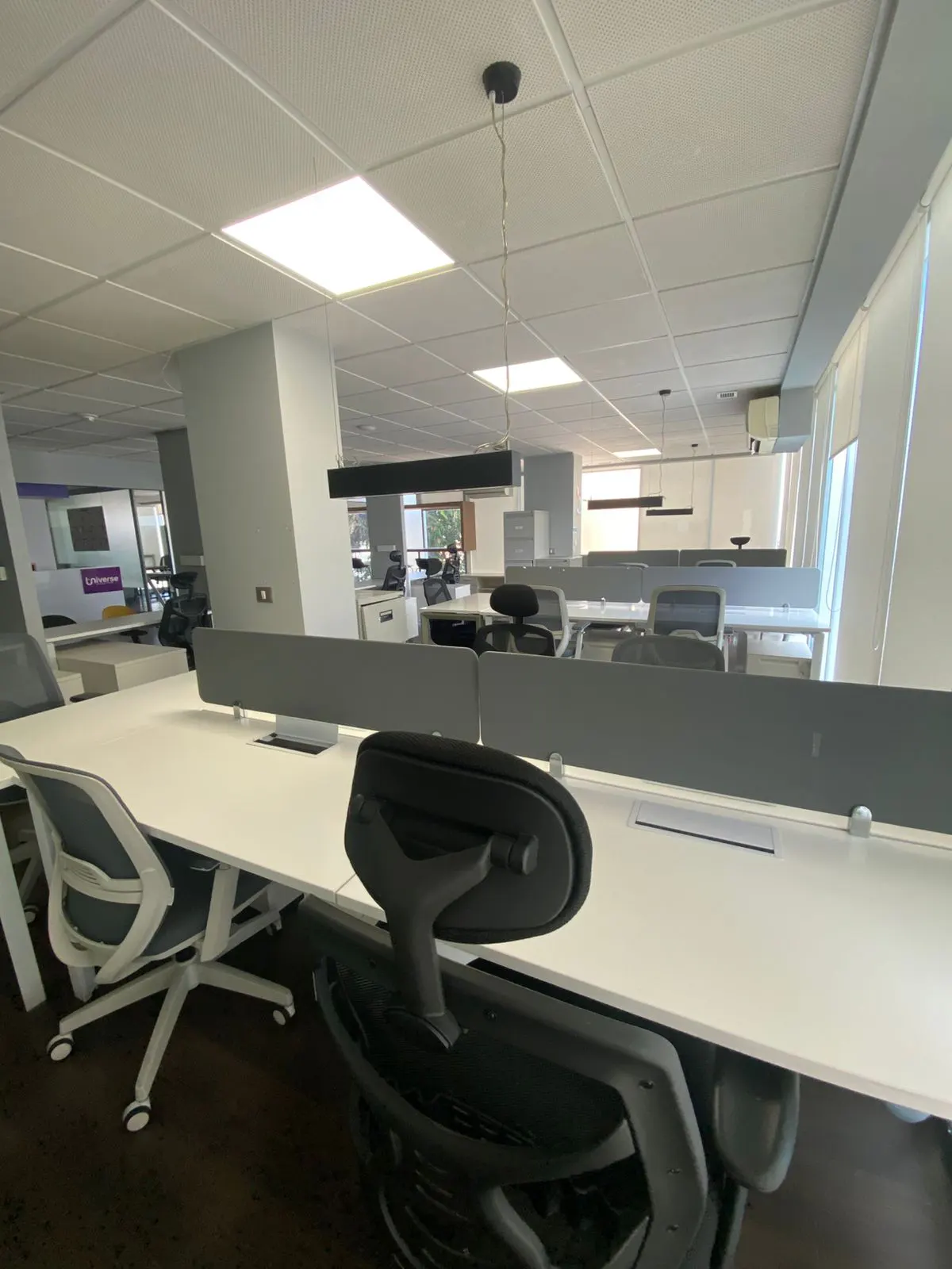 A bright, modern office space with white desks, gray partitions, and black chairs under fluorescent lighting.