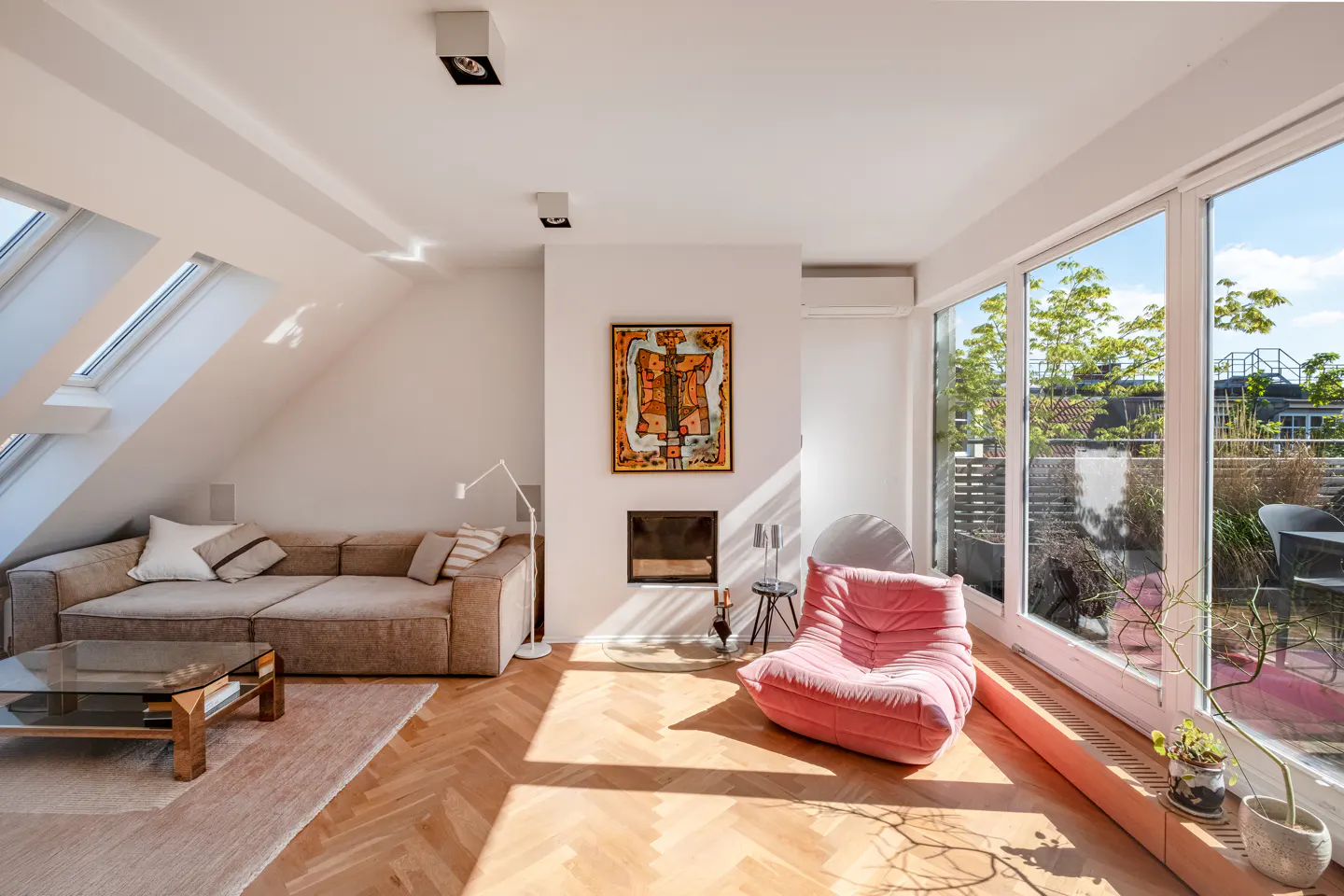 Bright living room with herringbone wood floors, a beige sofa, pink chair, and large windows overlooking a green outdoor space.