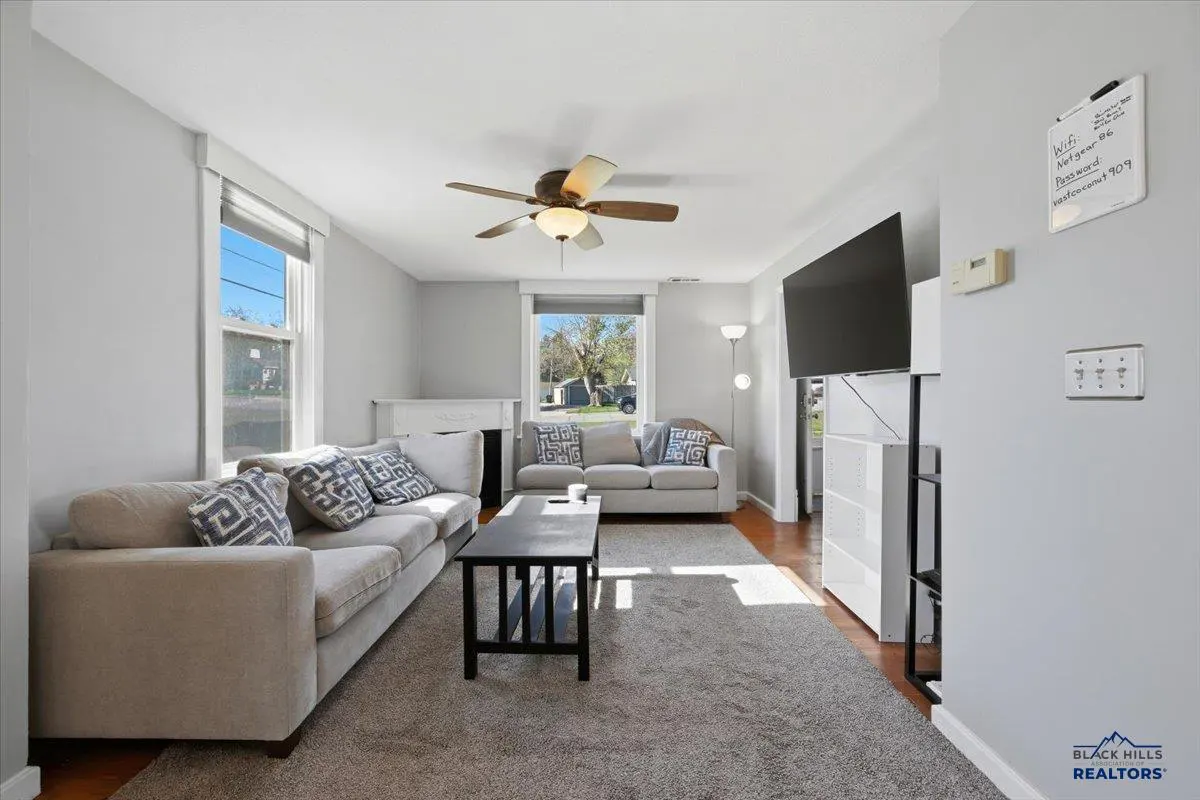 Living room with gray walls, a ceiling fan, and two beige sofas with patterned pillows. A black coffee table sits on a gray rug.