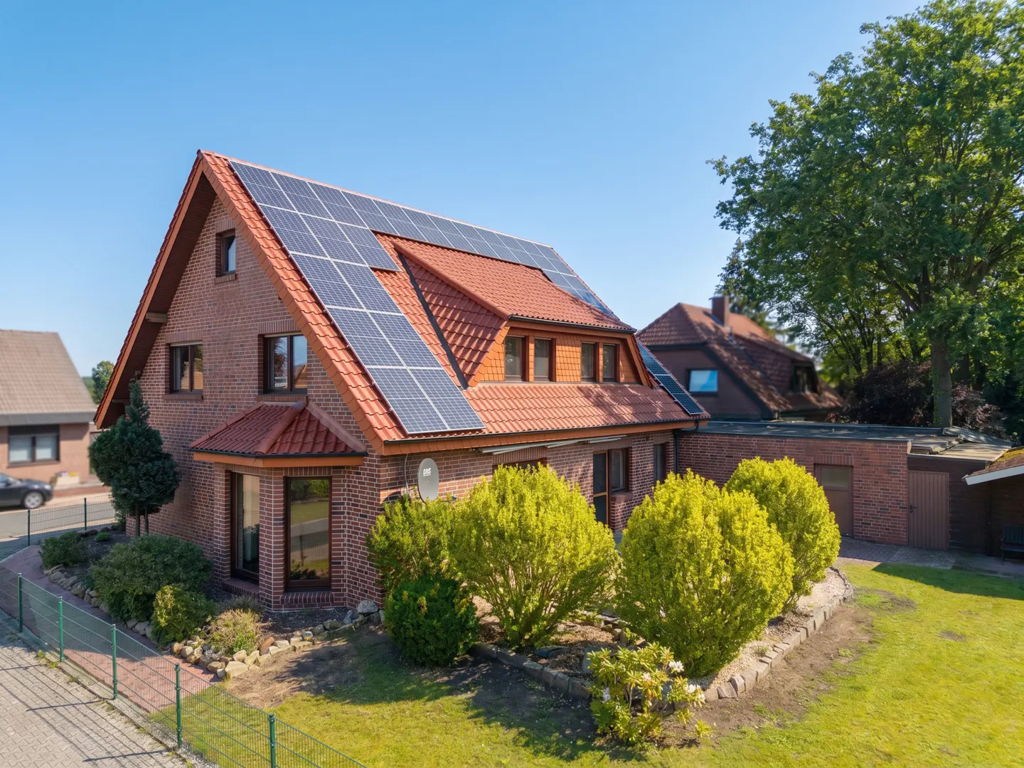 Two-story brick house with a red tile roof and solar panels. Green lawn and bushes surround the house. Blue sky.