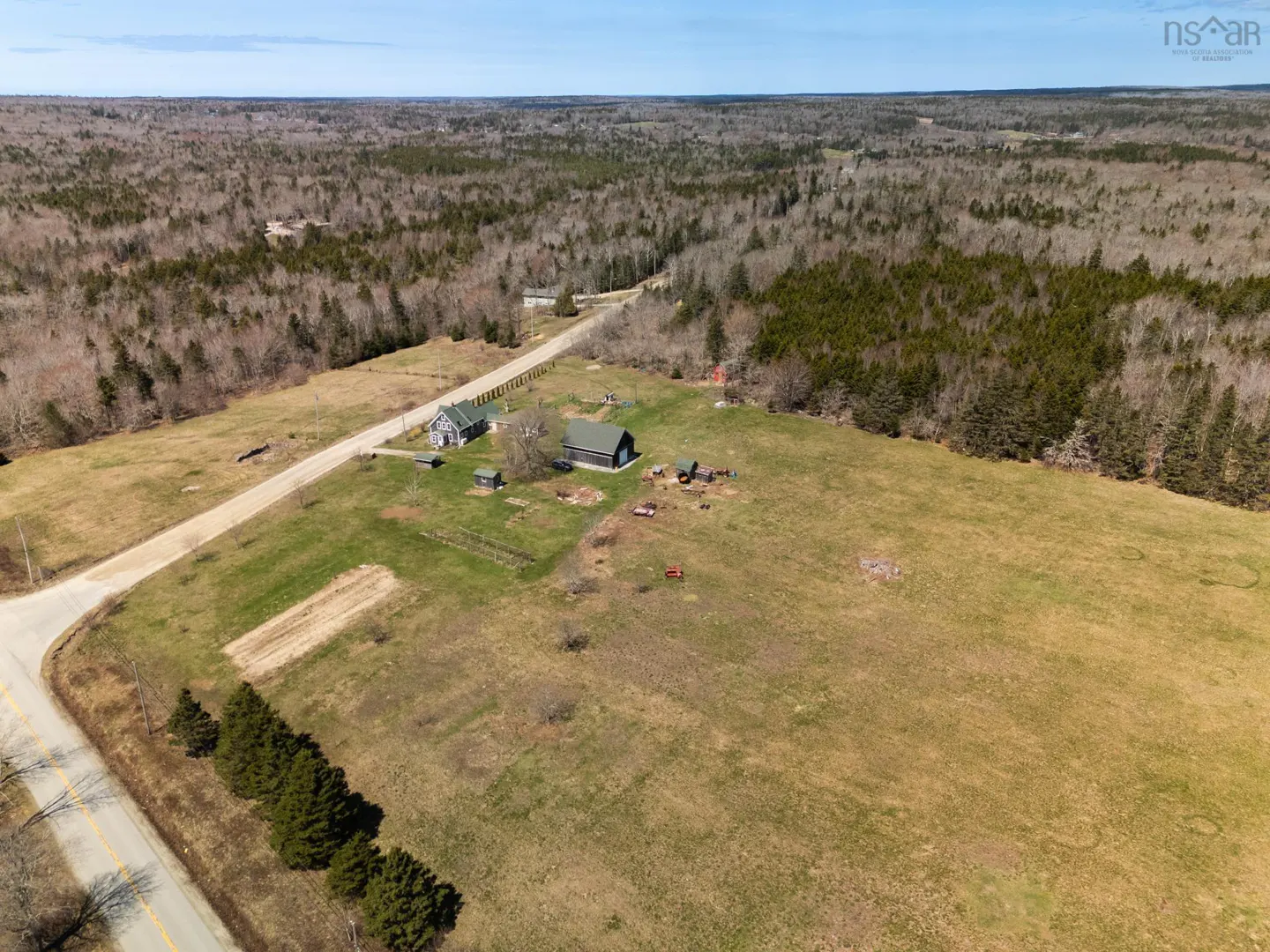 Aerial view of a rural property with a house, barn, and large grassy field bordered by trees and a road.