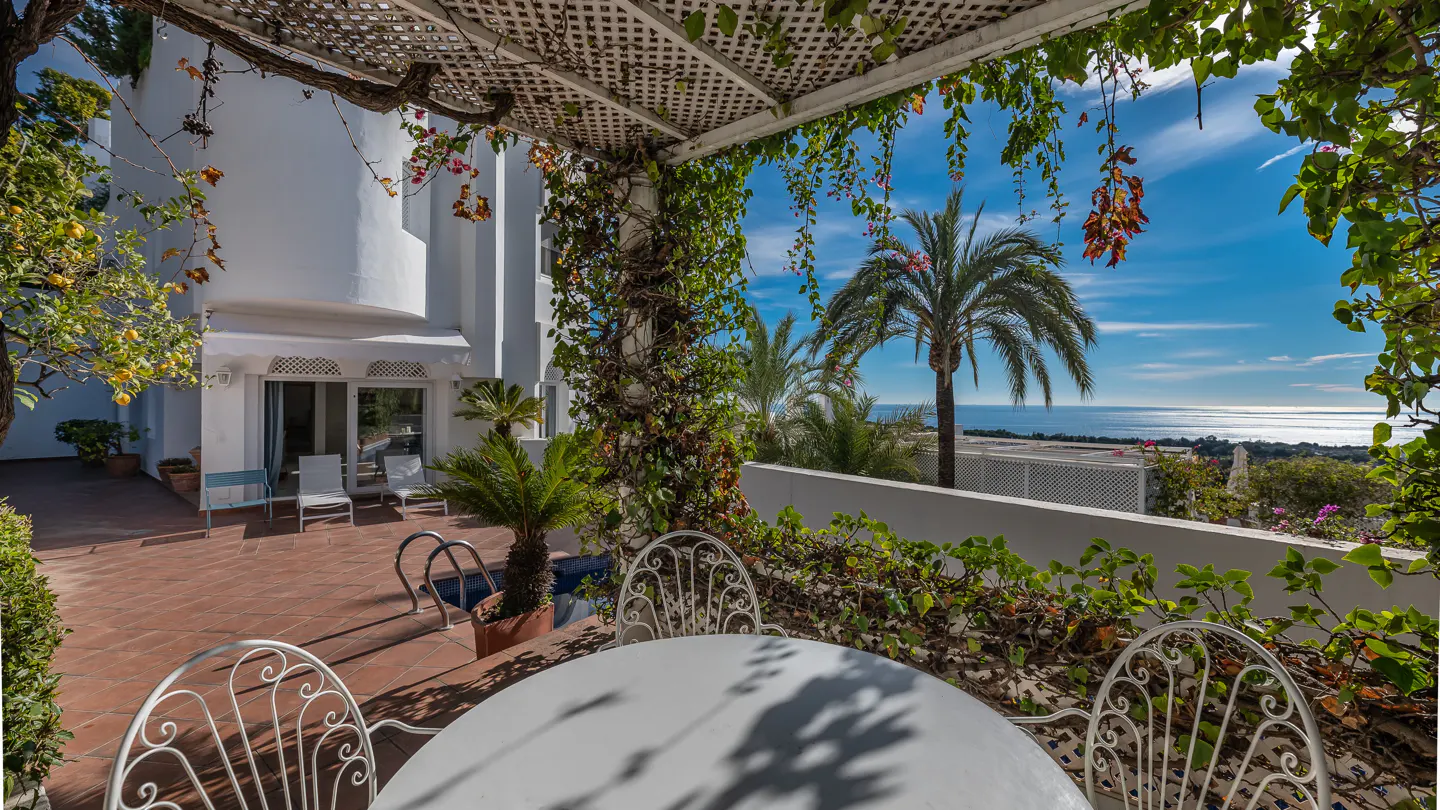 A white patio table and chairs under a vine-covered pergola overlooking a pool, white villa, and the blue ocean.