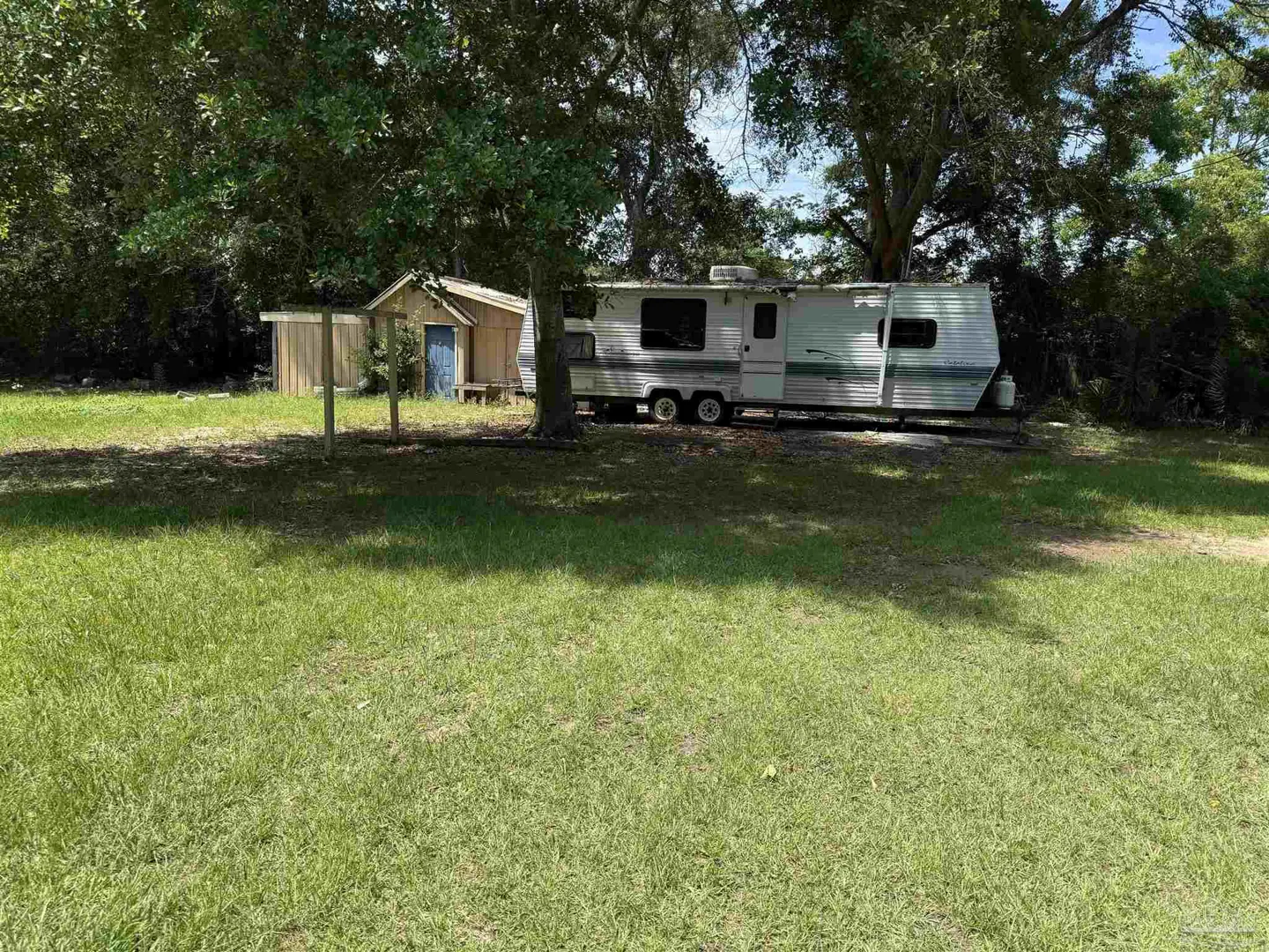 A white RV sits on a grassy lot next to a small wooden shed with a blue door. Trees surround the property.