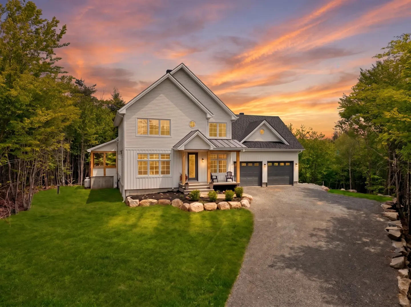 White two-story house with a gravel driveway and green lawn at sunset, surrounded by lush trees.