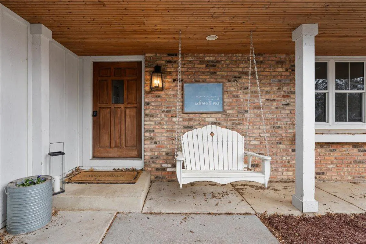 Covered porch with a wooden door, brick wall, and a white porch swing hanging from the wood ceiling.