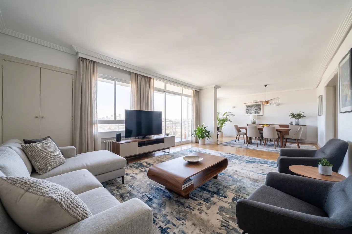 Bright, open-concept living and dining area with a gray sectional, armchairs, and a wood dining table. Large windows offer city views.