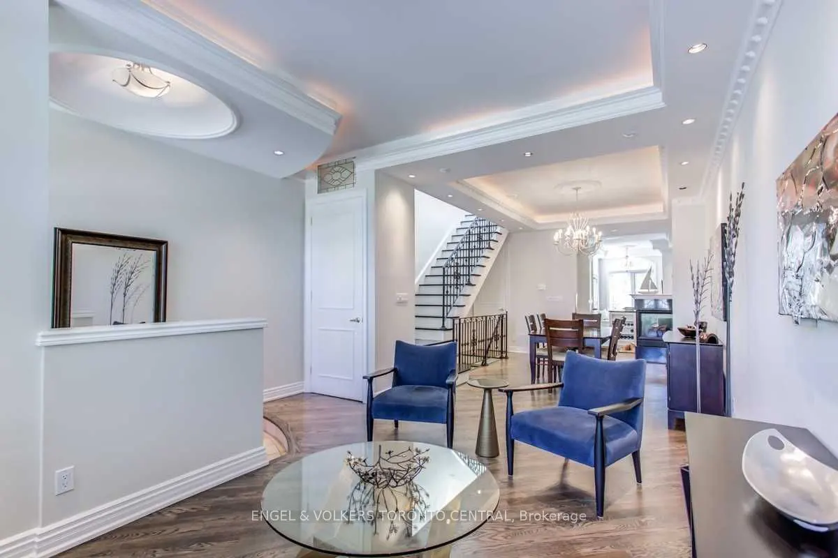 Bright, modern living room with blue chairs, glass table, and staircase in the background. White walls and dark wood floors.