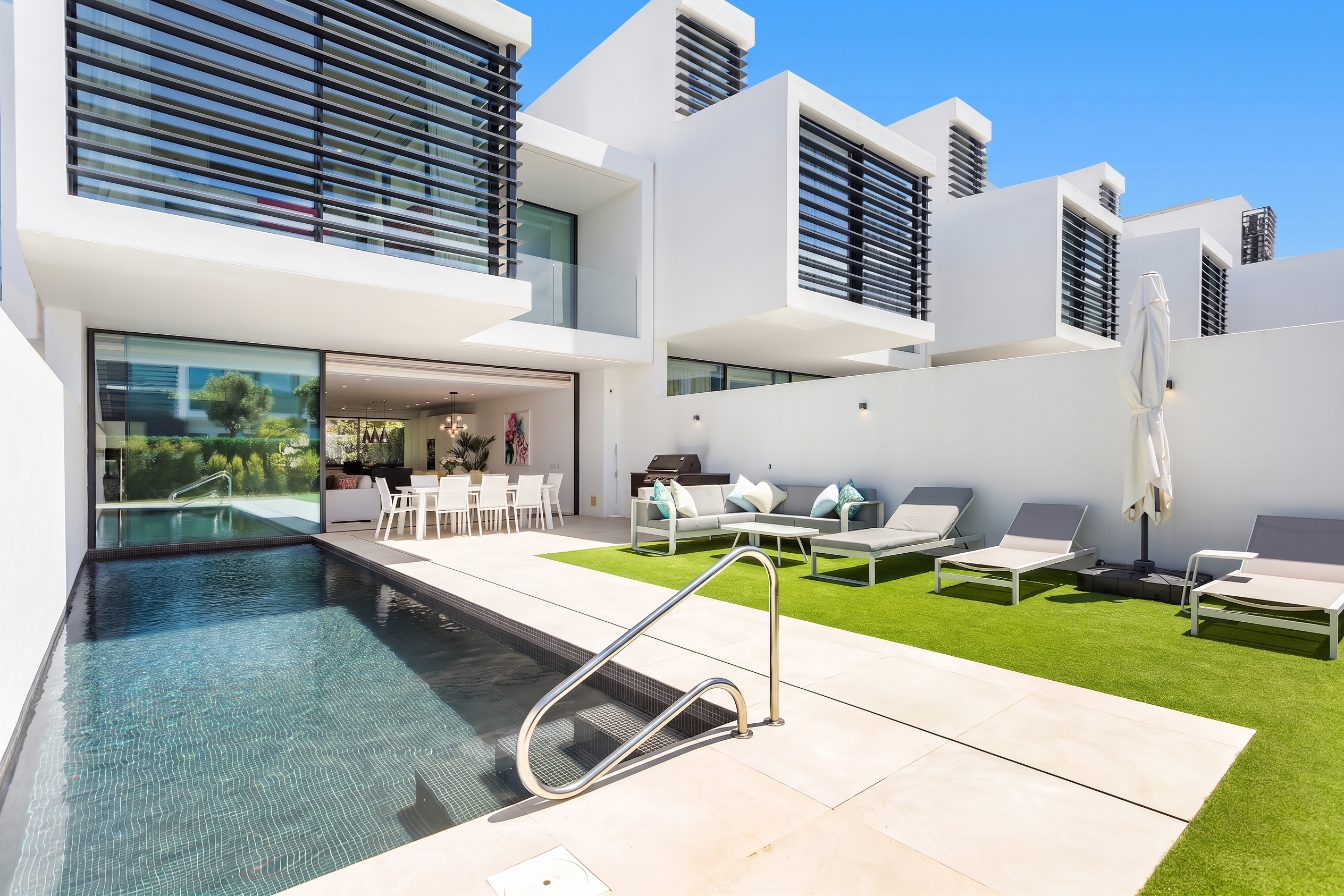 Modern white villa with a swimming pool, artificial grass lawn, lounge chairs, and an open-plan dining area.