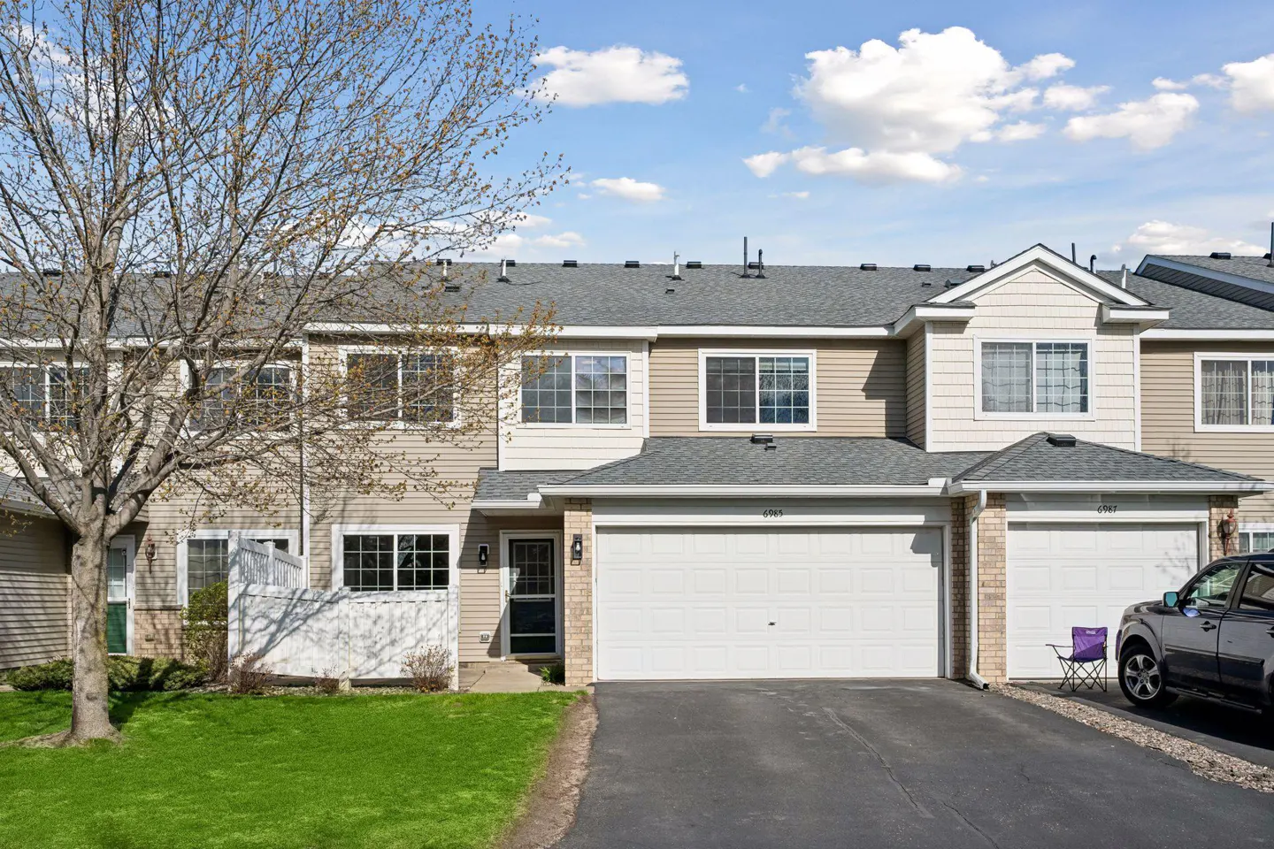 Tan townhouse with white garage doors, gray roof, and green lawn. A bare tree is on the left. A gray SUV is parked on the right.