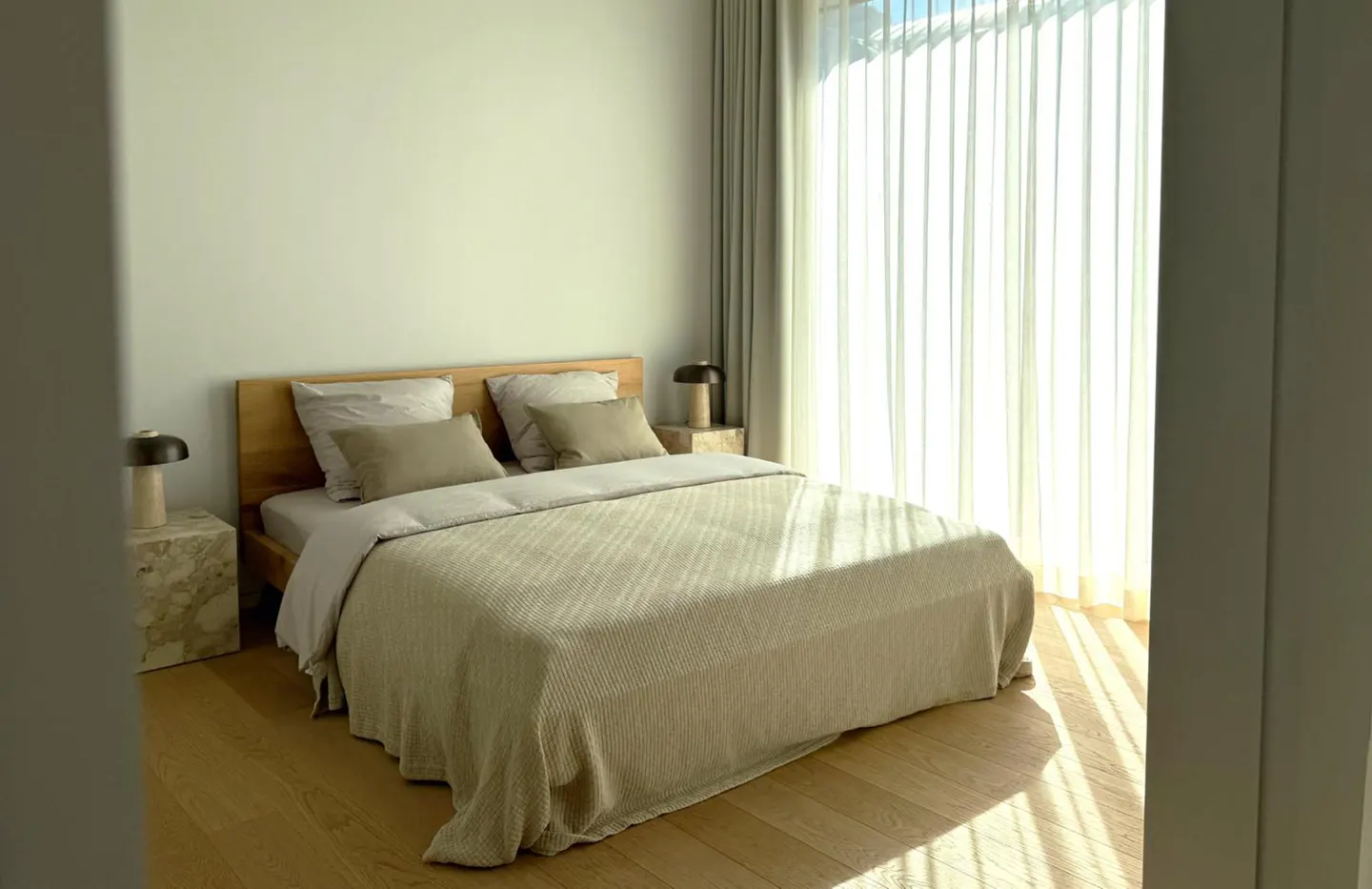 Bright bedroom with a wooden bed frame, beige bedding, and sheer white curtains letting in sunlight. Two marble nightstands with lamps are on either side.
