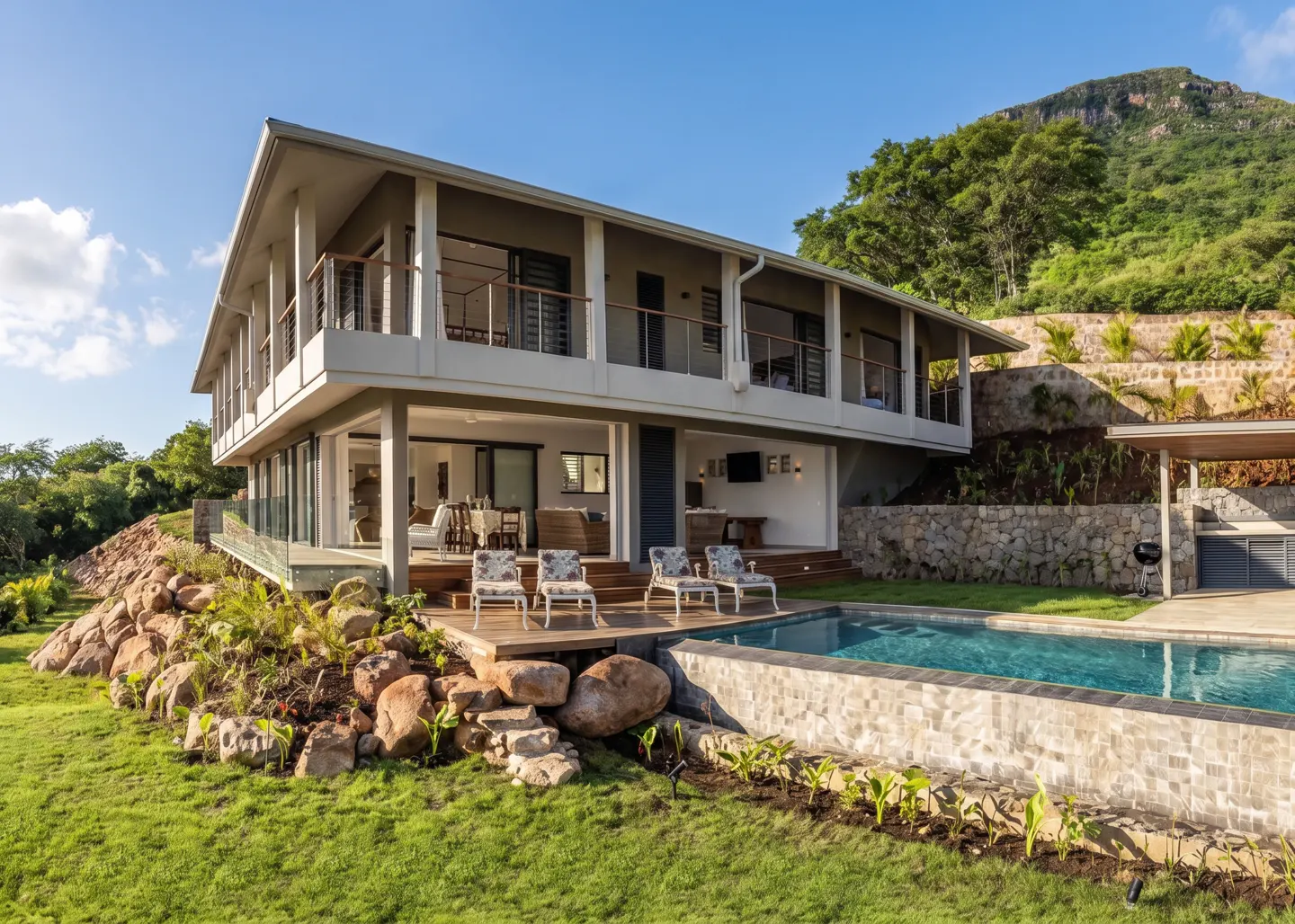 Two-story gray house with a pool, deck, and four chairs, set against a green hillside and blue sky.