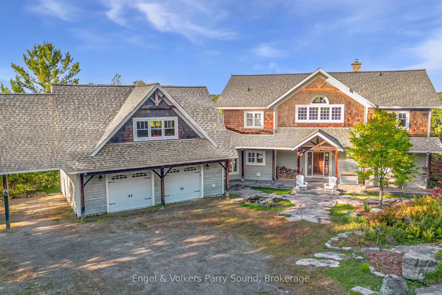 Exterior view of a two-story house with a two-car garage, gray siding, and a stone walkway. The house has a brown shingle roof and a covered porch with white chairs.
