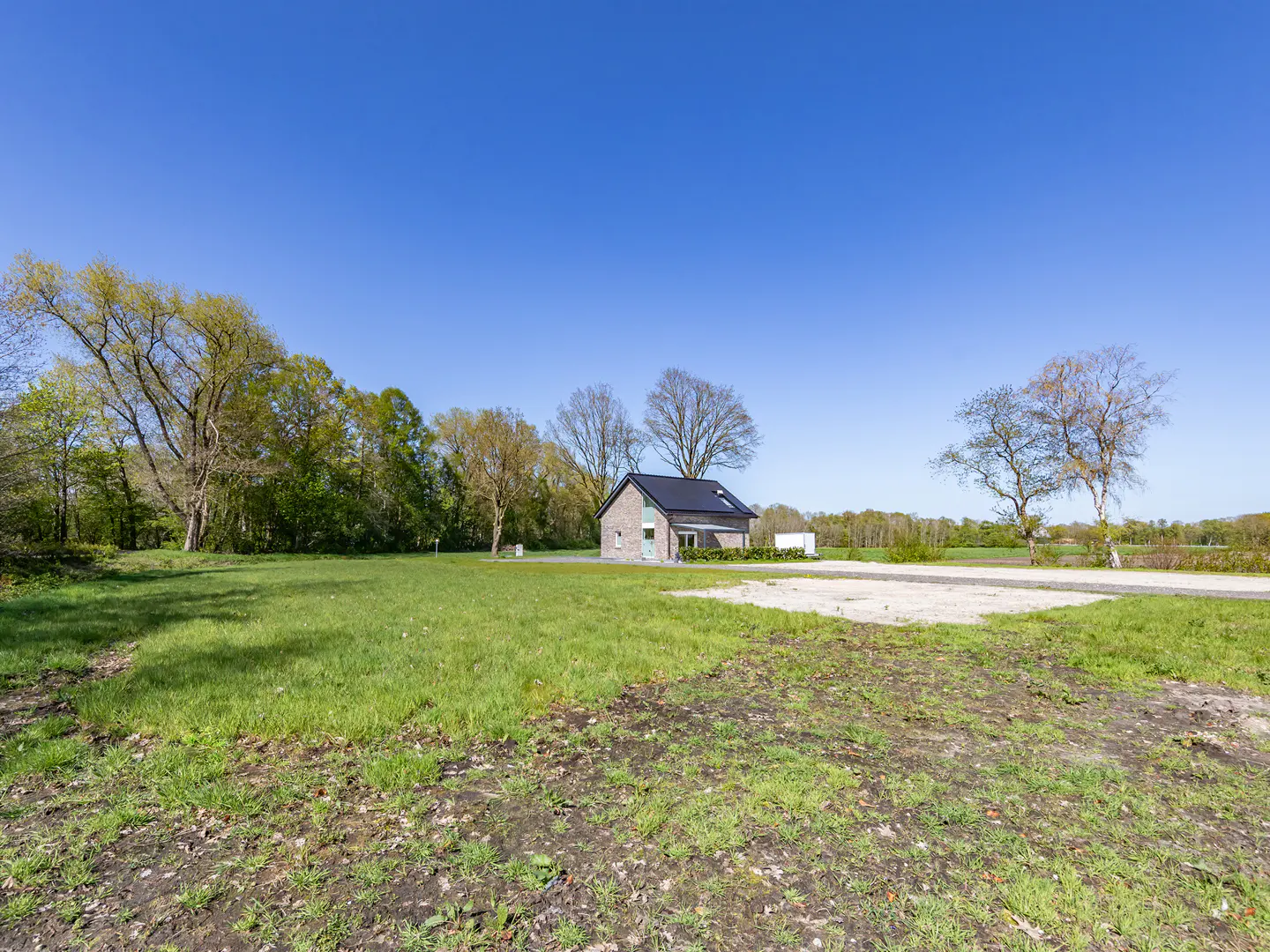 A single-story brick house with a black roof sits on a large grassy lot under a clear blue sky. Trees line the left side of the property.