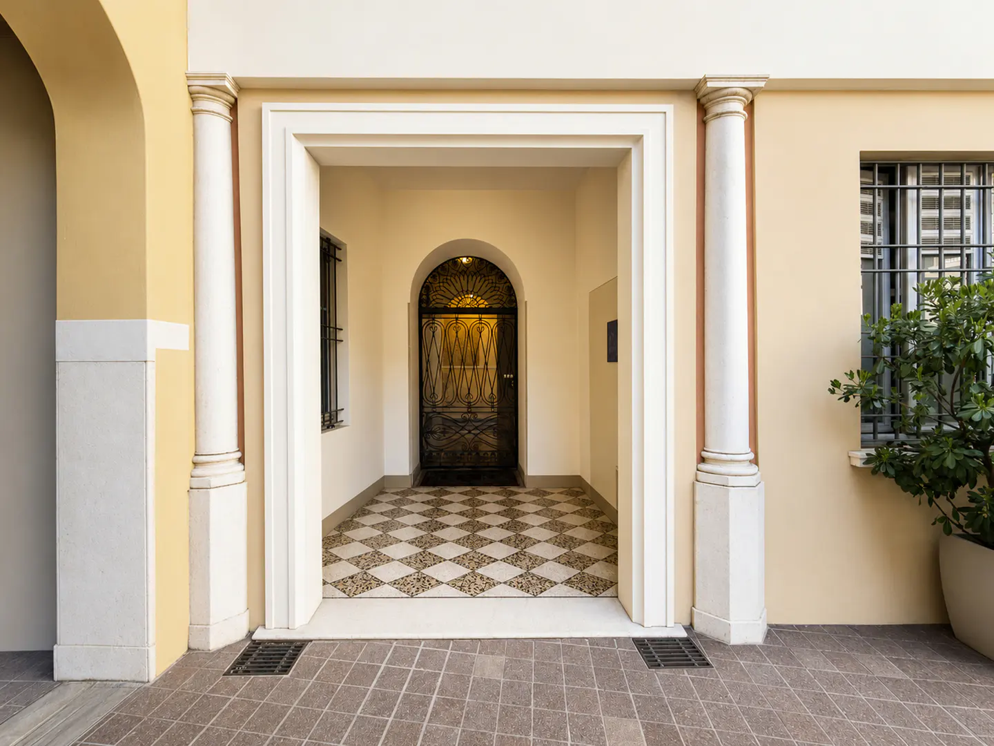 Entrance to a building with white columns, a black wrought iron door, and a diamond-patterned tile floor. A potted plant sits near a barred window.