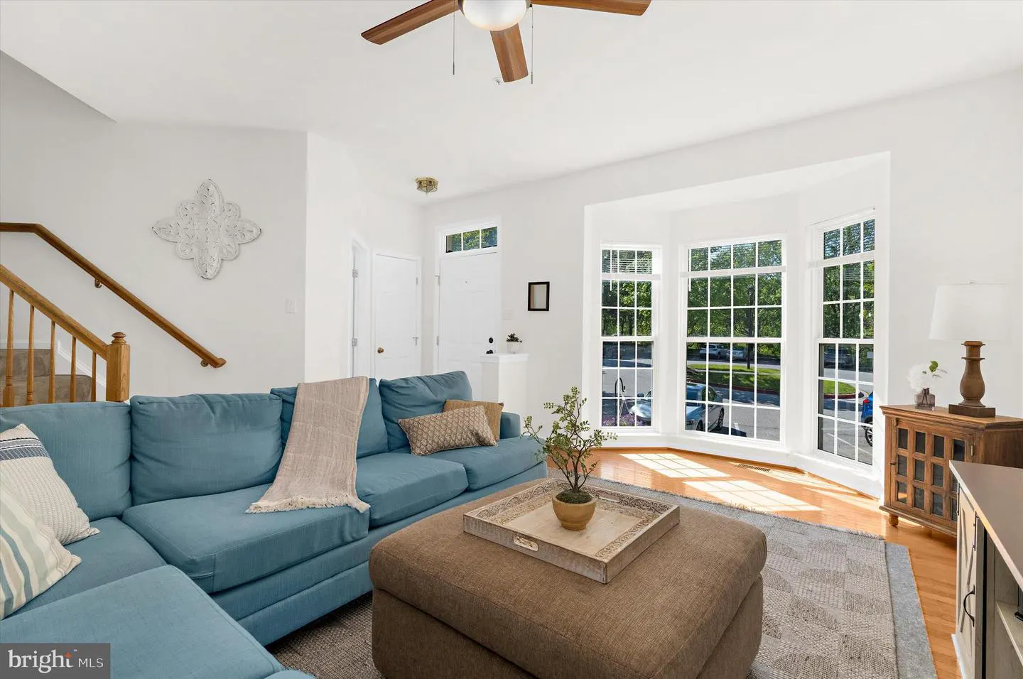 Bright living room with a blue sectional sofa, brown ottoman, and large bay windows. A wooden staircase is visible on the left.