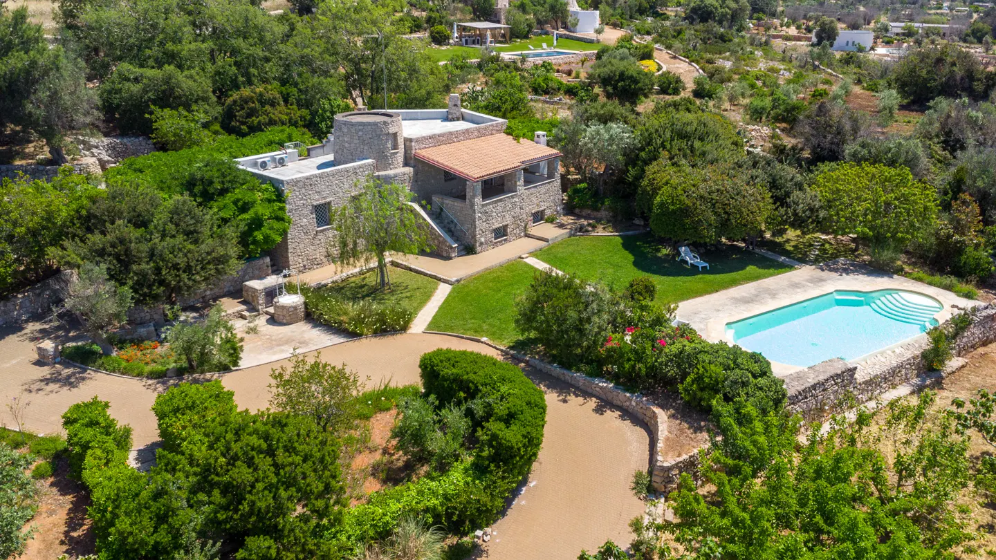 Aerial view of a stone villa with a round tower, swimming pool, and lush garden surrounded by trees.