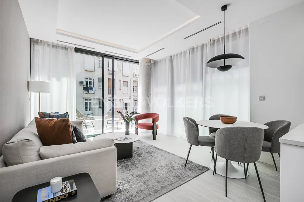 Bright living room with a gray sofa, red chair, round table with gray chairs, and a balcony with city views.