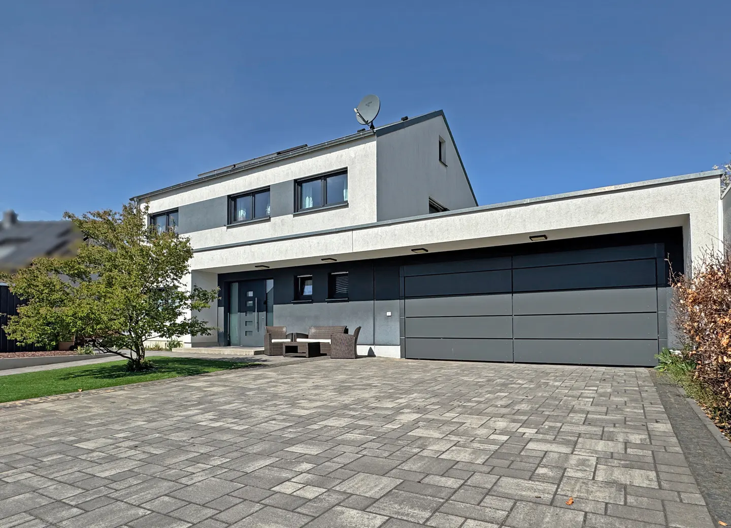 Modern two-story house with gray garage door, gray brick driveway, and outdoor seating. Blue sky.