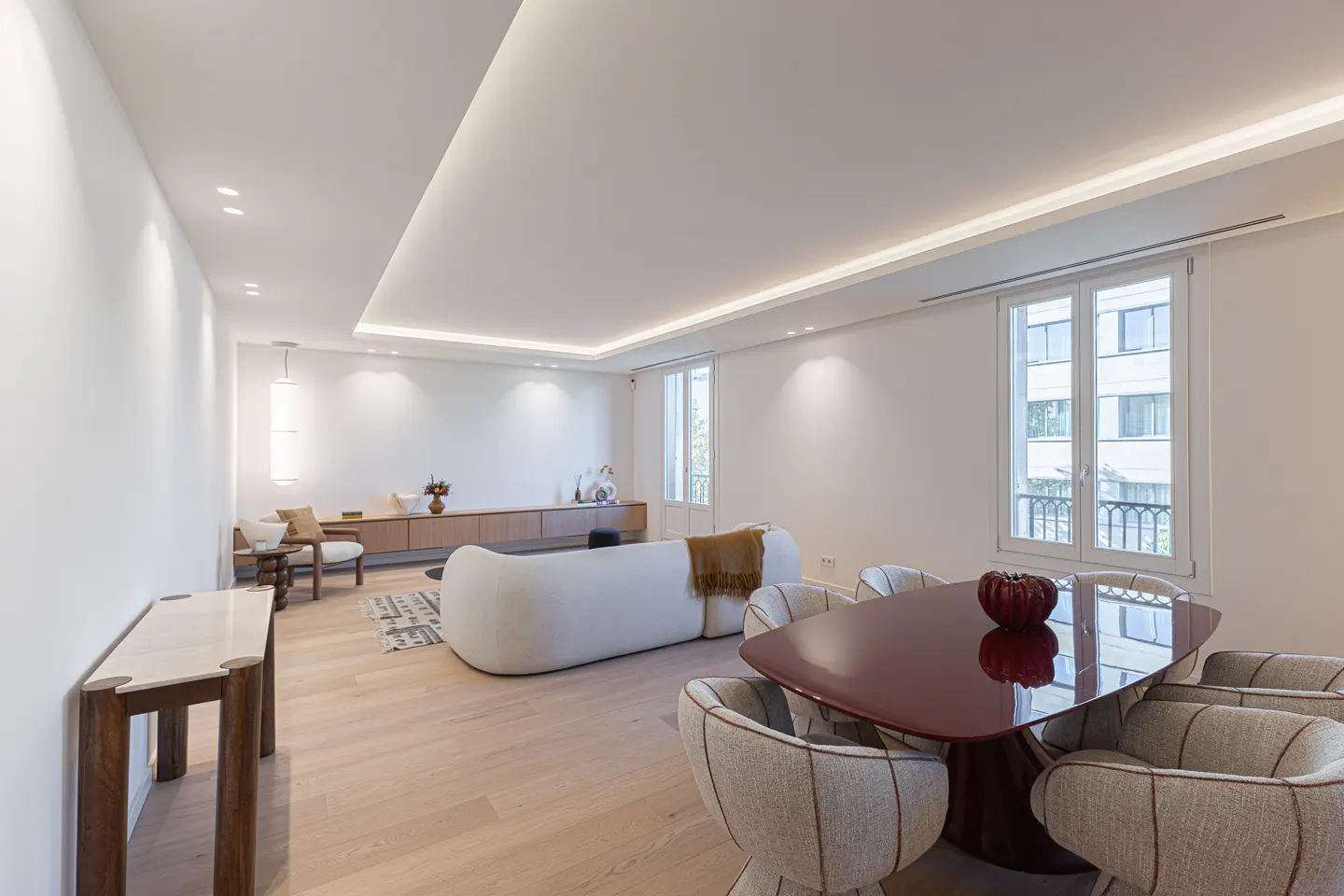 Bright, modern living and dining area with white walls, wood floors, and recessed lighting. A red table with six chairs sits near a window.