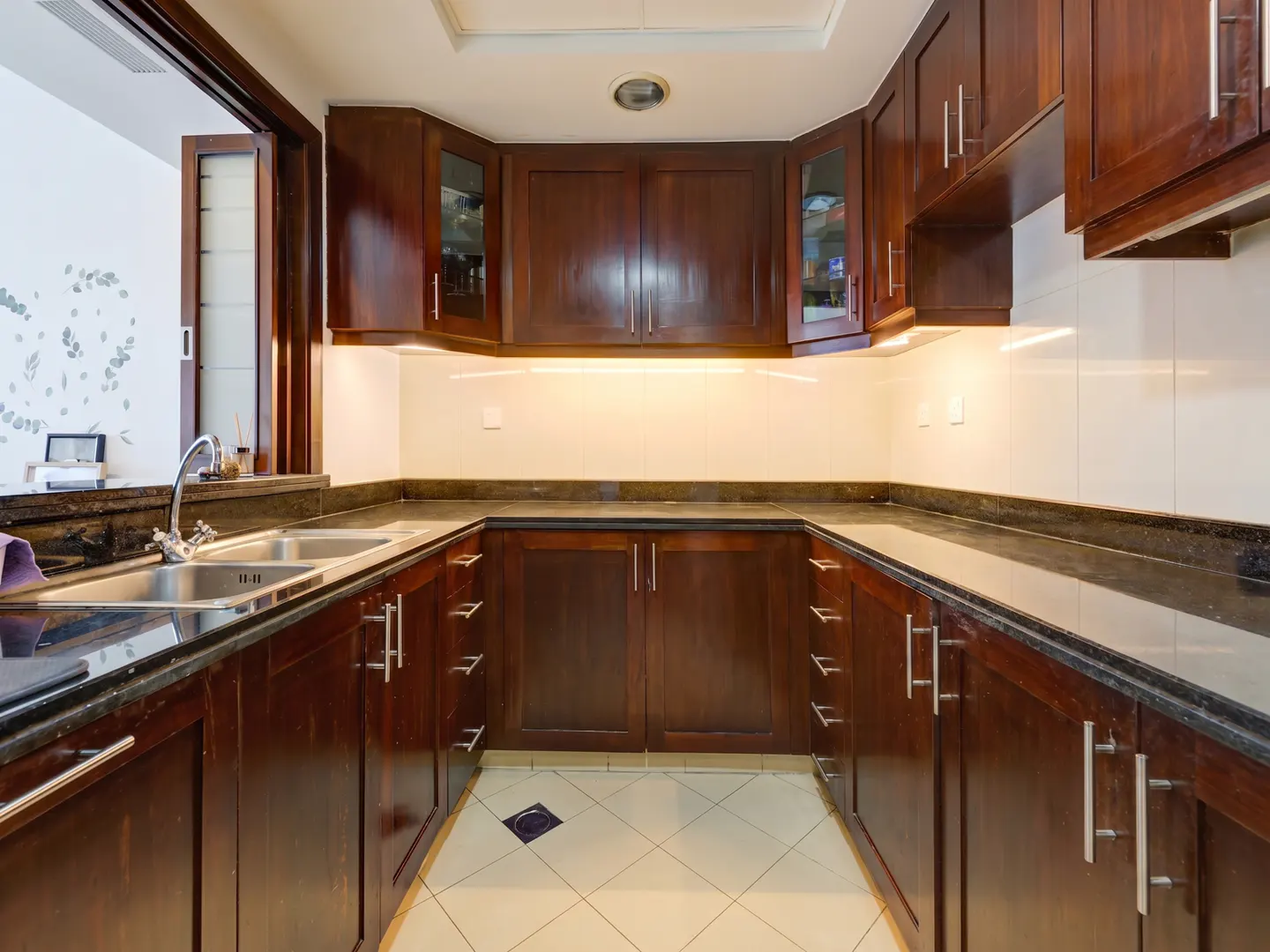A U-shaped kitchen with dark wood cabinets, black granite countertops, and white tile flooring. A window is on the left side of the image.