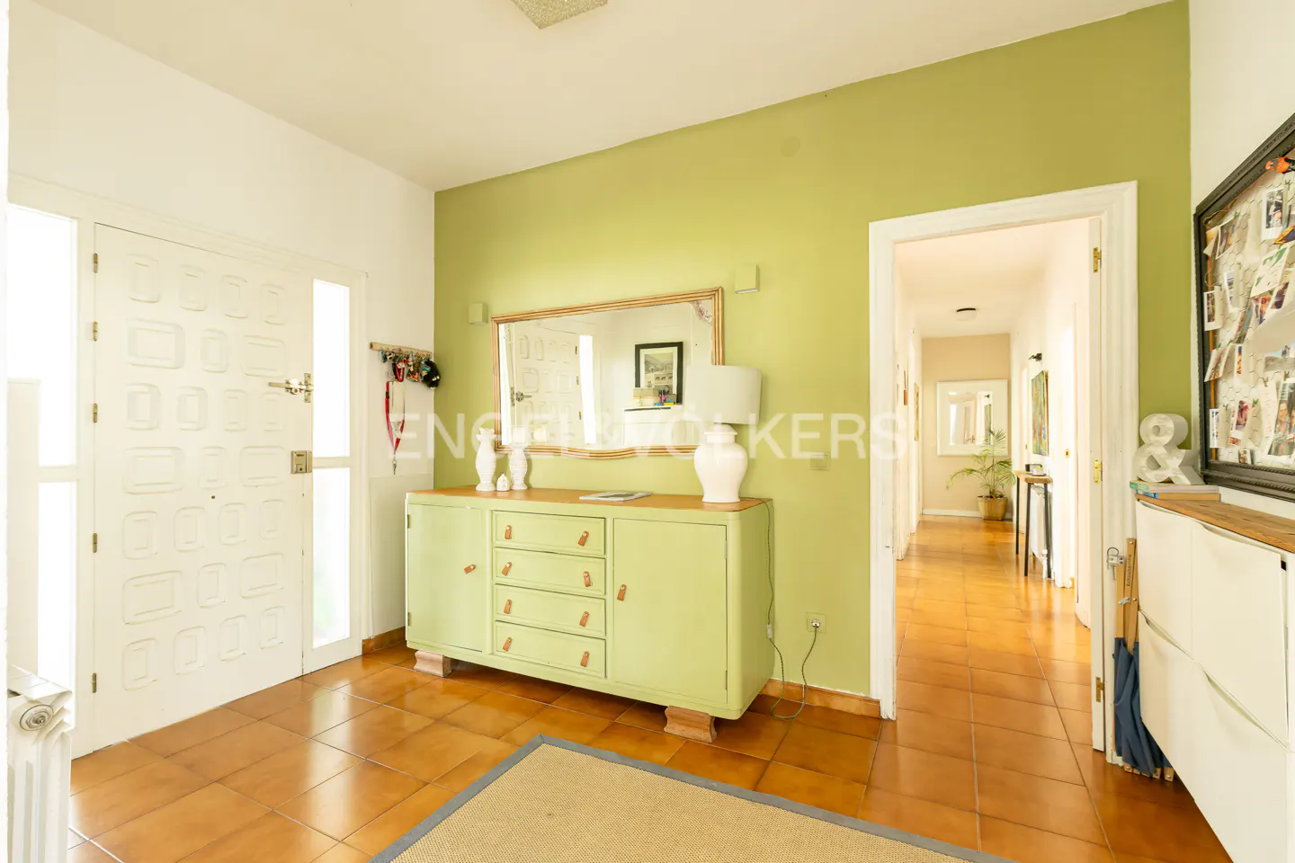 A home's entryway with a white door, green walls, and brown tile floors. A green cabinet sits beneath a mirror. A hallway is visible through a doorway.