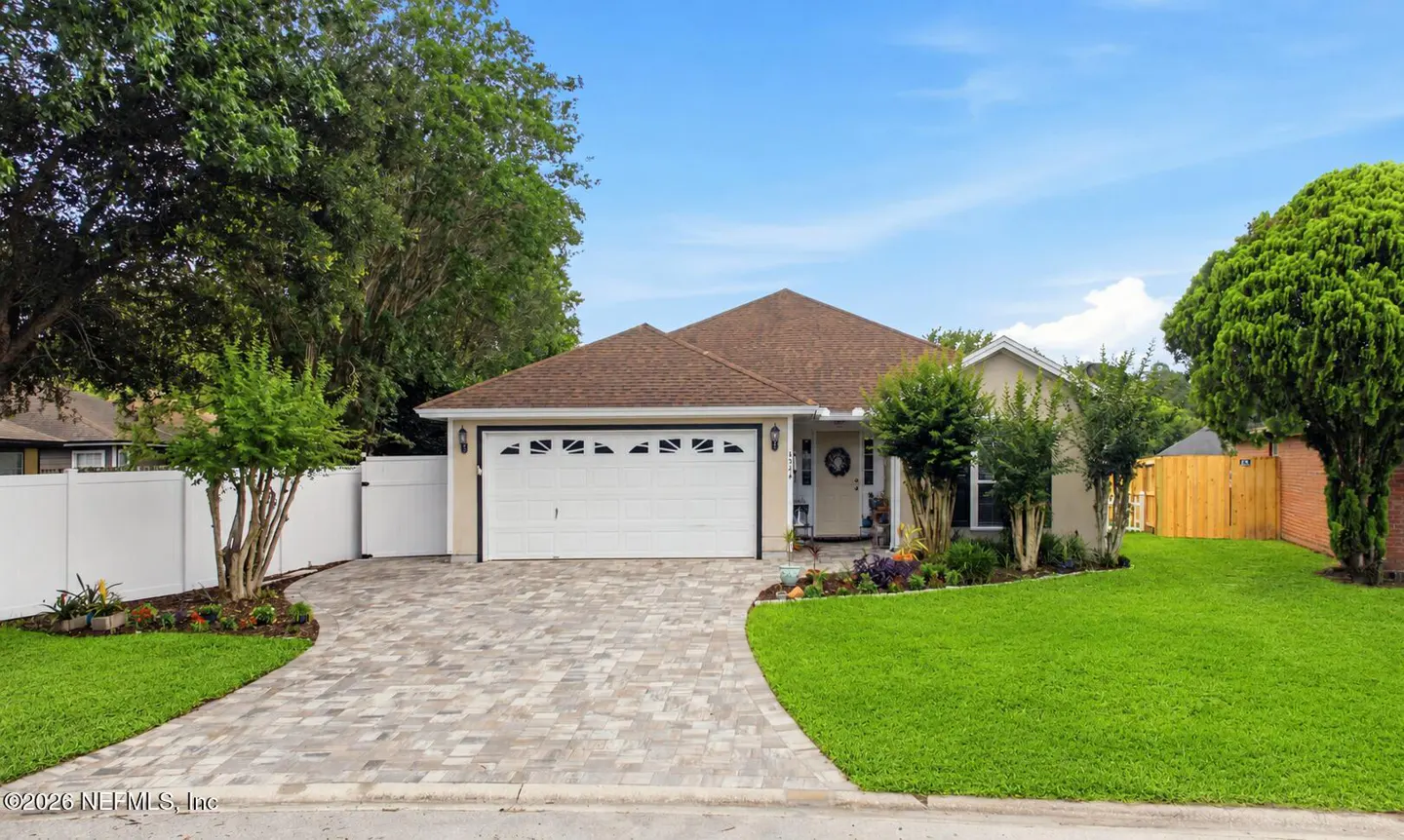 A single-story beige house with a brown roof, a white garage door, and a brick driveway. Green grass and trees surround the house.
