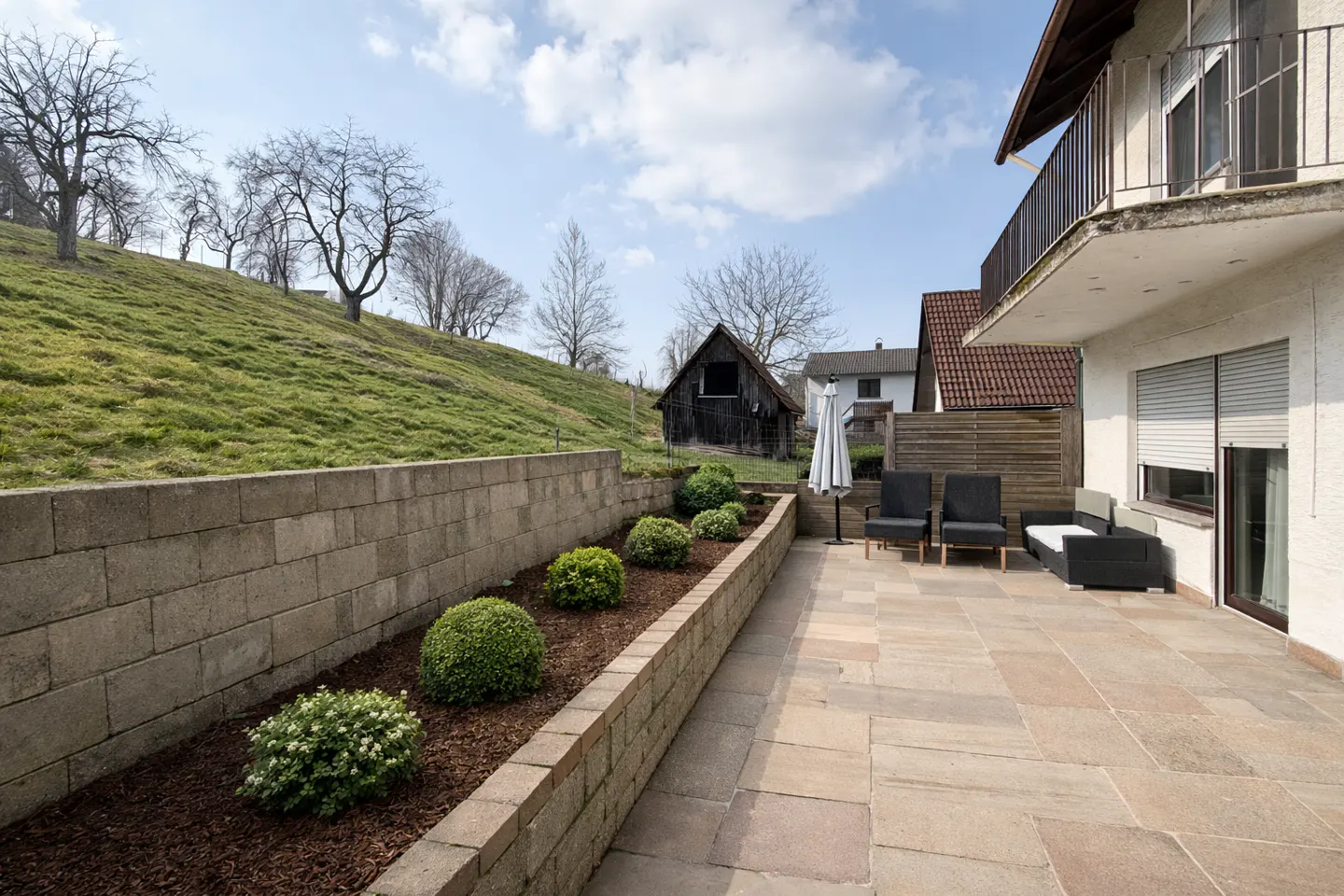 Stone patio with outdoor seating, a retaining wall with bushes, and a grassy hill in the background.