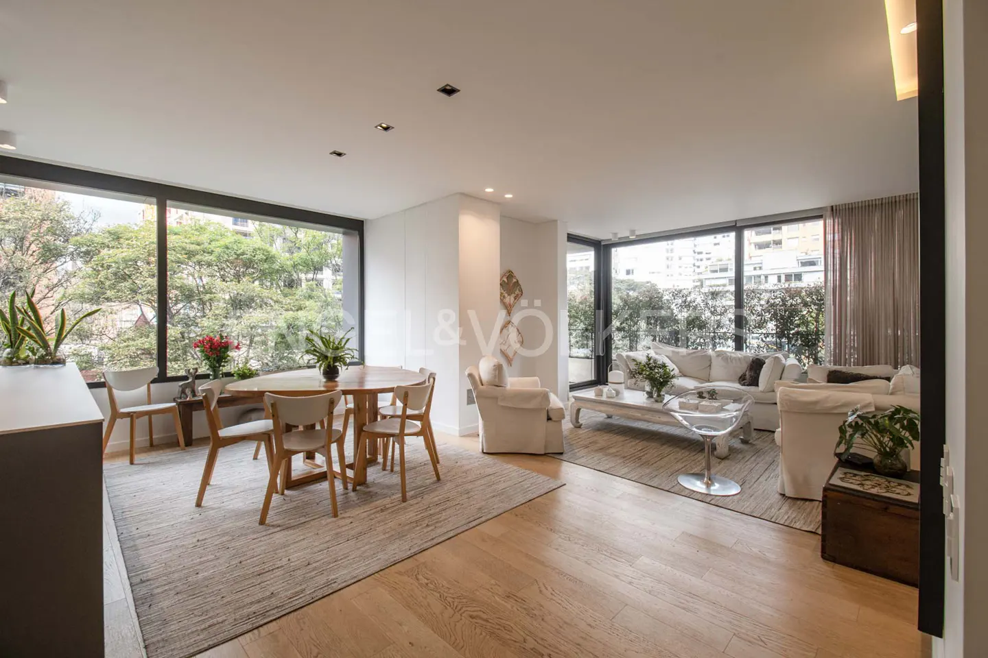 Bright, open-plan living and dining area with wood floors, large windows, and neutral decor. A round table and white sofas create a welcoming space.