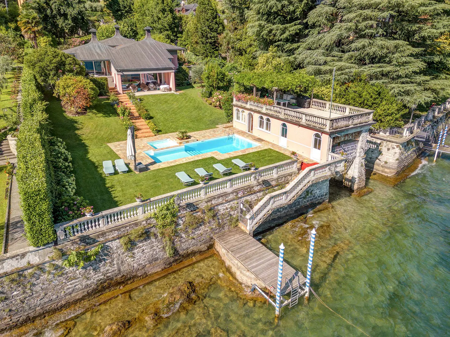 Aerial view of a pink lakefront villa with a pool, dock, and lush green landscaping on a sunny day.