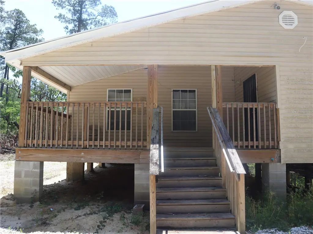 Tan house with a wooden porch and stairs leading up to the front door. The house is elevated on concrete blocks.